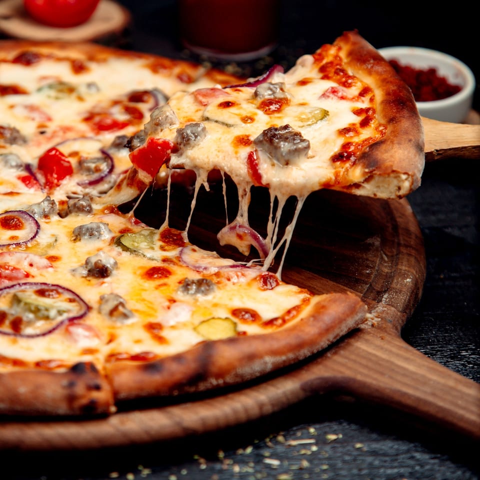 A slice of Wicked Pizza Company's pizza with melted cheese, meat, and vegetables being lifted.